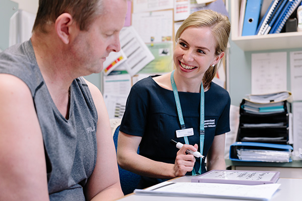 A woman in a hospital uniform sits beside a man at a desk in an office.