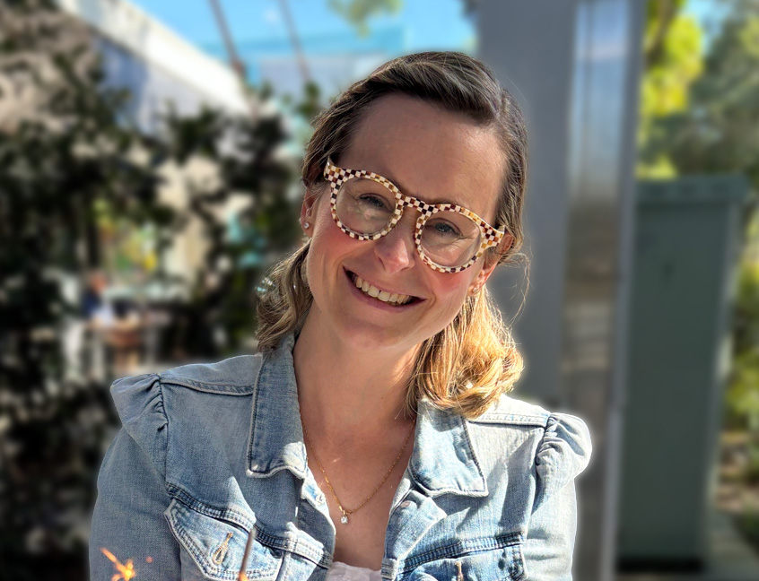 A woman with glasses wearing a denim jacket, smiling while standing outdoors.