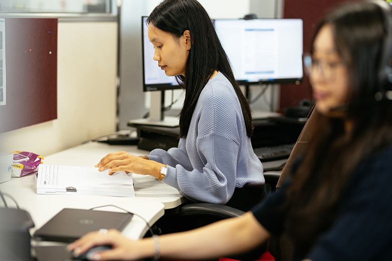 A woman seated at a desk, focused on her laptop while working in a well-lit office environment.