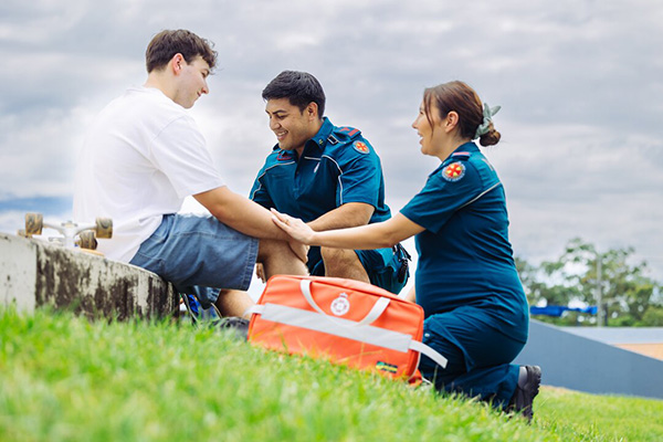 A paramedic assists a patient lying on the grass, providing medical care in an outdoor setting.