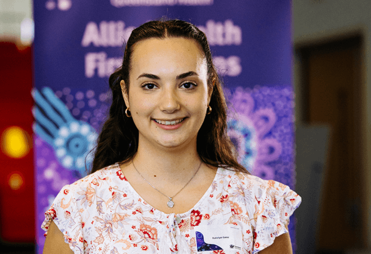 A young woman in a floral shirt stands in front of a colorful banner, smiling and engaging with the audience.
