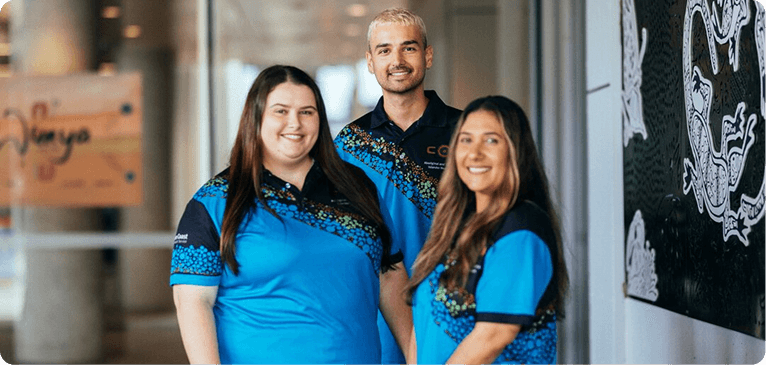 Three individuals in blue shirts stand together in front of a plain wall.