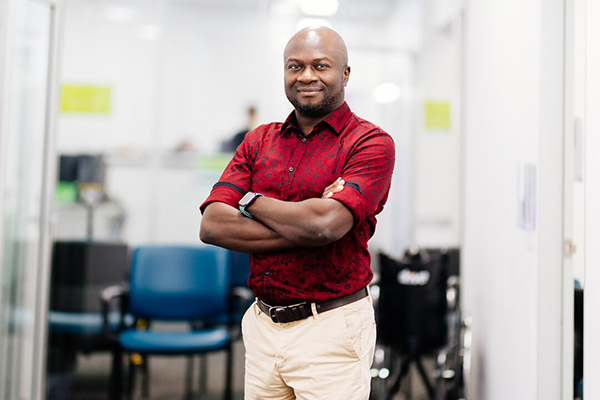 A man wearing a red shirt and tan pants stands in an office setting, looking towards the camera.