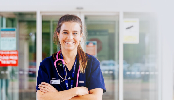 A young female doctor smiling and standing in front of a hospital.