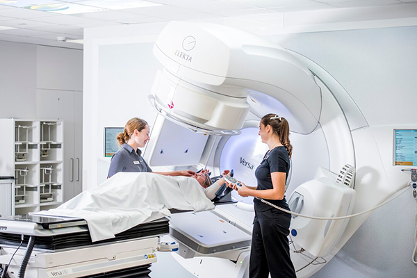 A man and woman stand in a hospital room beside an MRI machine, discussing the procedure in a clinical setting.