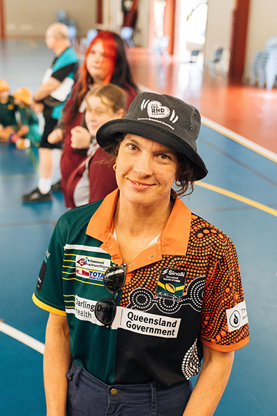A woman in a hat and shirt standing in a gym with children standing and sitting in the background.
