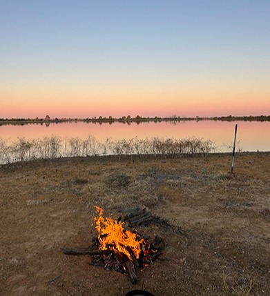 A campfire burns brightly on the shore of a tranquil lake, surrounded by rocks and trees under a clear sky.