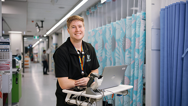 A man stands in a hospital hallway, focused on his laptop while surrounded by medical equipment and signage.