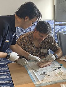 Two men in blue shirts and white gloves collaborate at a table, focused on their task.