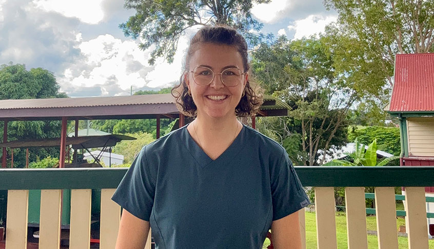 A woman in scrubs stands on a porch, smiling and looking towards the camera, with a welcoming expression.