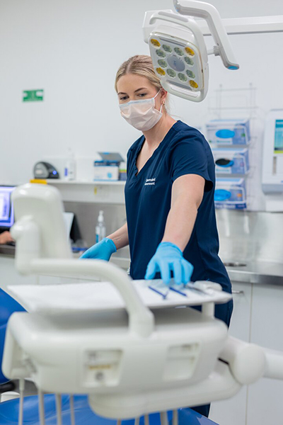A woman in blue scrubs reaching for a dental tool, next to a dental chair.