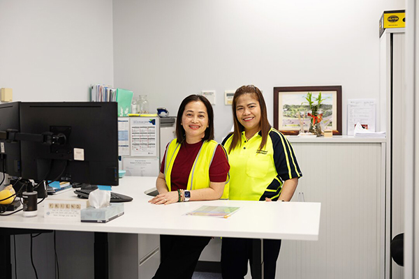 Two women in yellow vests standing at a desk, engaged in conversation and reviewing documents.