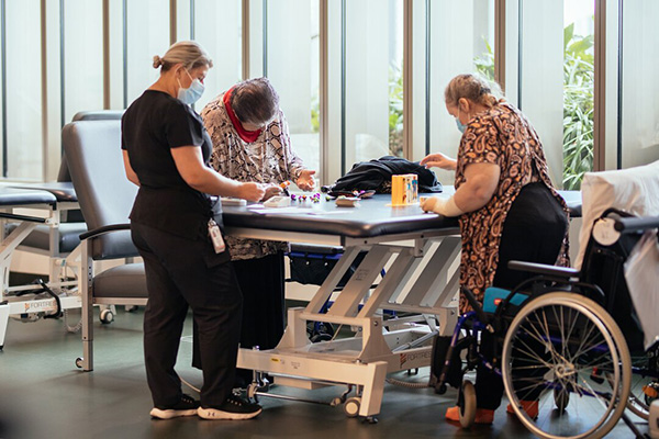 Two women leaning over a table doing motor skills activities while a woman in scrubs observes them.