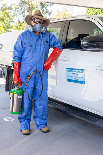 A man in a blue coverall wearing a mask stands beside a white truck, holding a spray container.