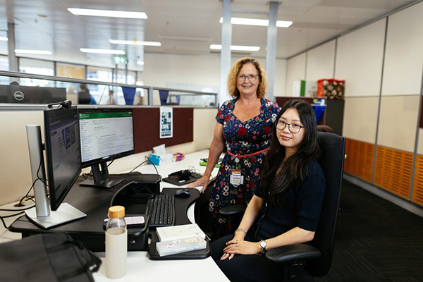Two women engaged in conversation while one woman is sitting at a desk with computer screens.