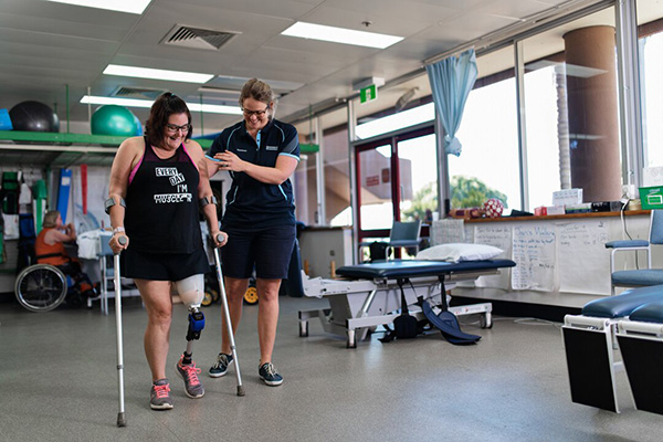 A physiotherapist next to a women with a prostetic leg and on crutches teaching her walk again.