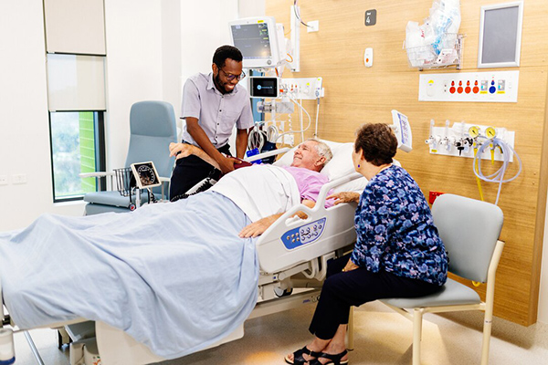 A man lying in a hospital bed with a man in a hospital uniform taking his blood pressure, and a woman sitting beside him and .