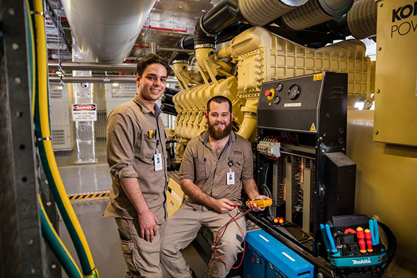 Two men in work clothes stand beside a generator, discussing its operation and maintenance.