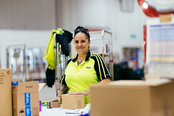A woman wearing a yellow shirt stands in a spacious warehouse, surrounded by shelves and boxes.