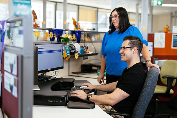 A man seated at a desk, focused on his work at a computer, with a woman standing beside him.