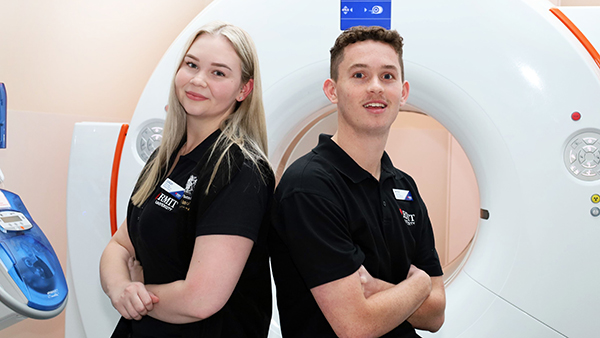 Two individuals stand in front of an MRI machine, discussing its features and operation in a medical setting.